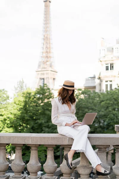 Mujer con estilo en sombrero de sol usando el ordenador portátil en la calle urbana con la torre Eiffel en el fondo en París - foto de stock