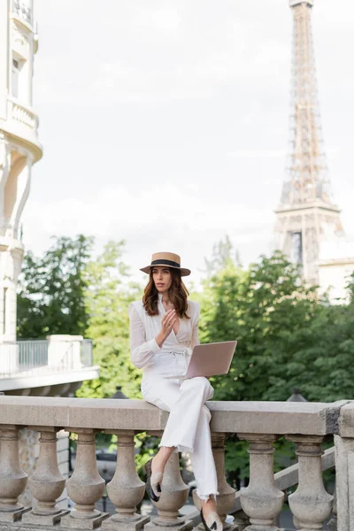 Mujer de moda en sombrero de sol que sostiene el ordenador portátil en la calle con la torre Eiffel en el fondo en París - foto de stock