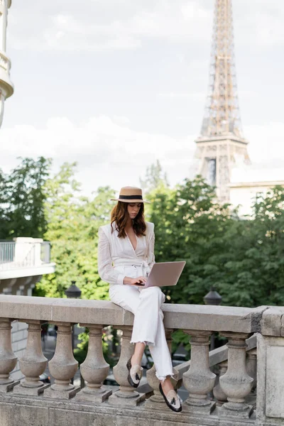 Jeune femme élégante utilisant un ordinateur portable dans la rue urbaine avec la tour Eiffel floue à l'arrière-plan à Paris — Photo de stock