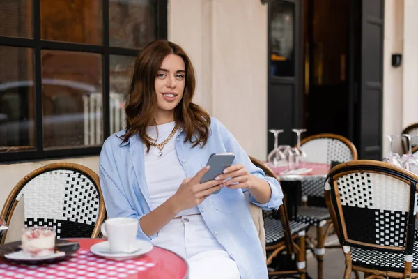 Mujer sonriente usando teléfono inteligente cerca de postre y café en la terraza de la cafetería en París - foto de stock