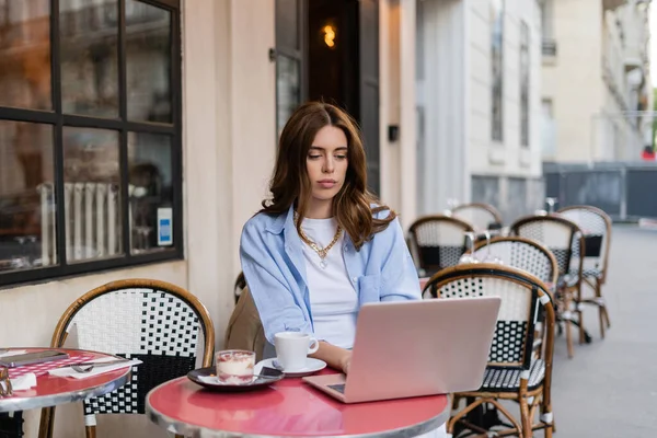 Élégant pigiste utilisant un ordinateur portable près du café et du dessert sur la terrasse du café à Paris — Photo de stock