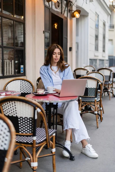 Jeune pigiste utilisant un ordinateur portable près du dessert et du café sur la terrasse du café à Paris — Photo de stock