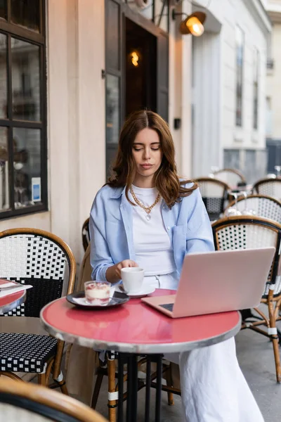 Freelancer holding coffee cup near dessert and laptop on table of cafe in Paris — Stock Photo