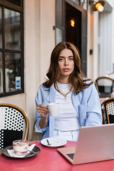Stilvolle Frau hält Tasse Kaffee in der Nähe verschwommenes Dessert und Laptop in Outdoor-Café in Paris — Stockfoto