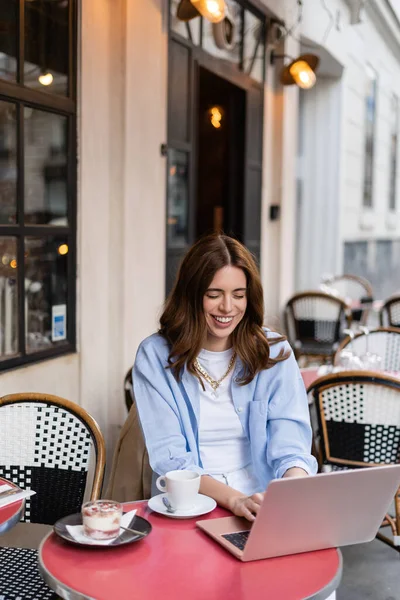 Fröhliche Freiberuflerin mit Laptop in der Nähe von Dessert und Kaffee in Outdoor-Café in Frankreich — Stockfoto