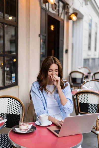 Positive freelancer using laptop near coffee and dessert in cafe on urban street in France — Stock Photo