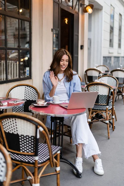 Femme souriante ayant un appel vidéo sur un ordinateur portable près du café dans un café extérieur en France — Photo de stock