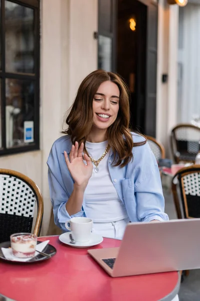 Femme élégante ayant un appel vidéo sur ordinateur portable près du café et du désert dans un café en plein air à Paris — Photo de stock