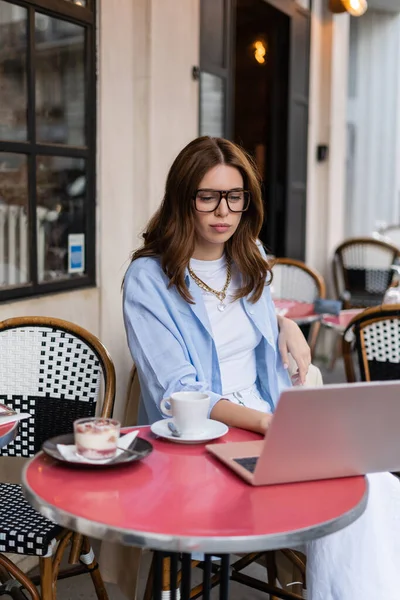 Stylish freelancer, використовуючи ноутбук біля десерту і каву на відкритому кафе в Парижі. — стокове фото