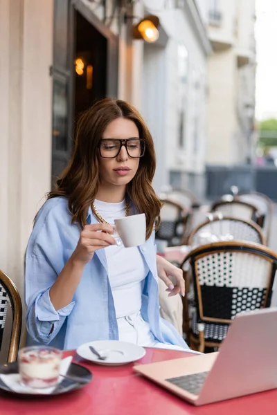 Femme élégante tenant une tasse près d'un ordinateur portable et dessert flou sur la terrasse du café à Paris — Photo de stock