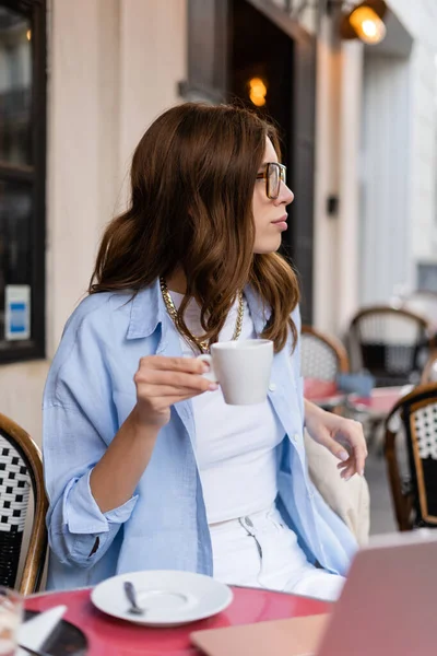 Vista lateral de la mujer joven de moda sosteniendo la taza de café cerca de la computadora portátil en la terraza de la cafetería en París - foto de stock