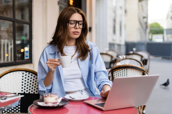 Модный фрилансер держит чашку возле ноутбука и десерт в открытом кафе в Париже — стоковое фото