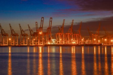 A night view of Khorfakkan Port in the United Arab Emirates ,showcases the lit up port facilities. The illuminated cranes and containers create a modern and busy atmosphere, while the dark waters of the sea provide a contrasting background.