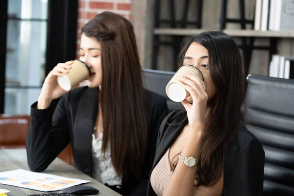 Office coffee break with two female colleagues sitting chatting over cups of coffee. Asian business woman holding coffee cup in office.