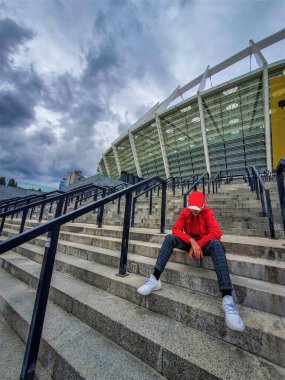 a young man in a red cap and red jacket sits on the stairs in front of the football stadium