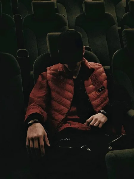 a guy in a red jacket and a black cap in a leather armchair in a movie hall.