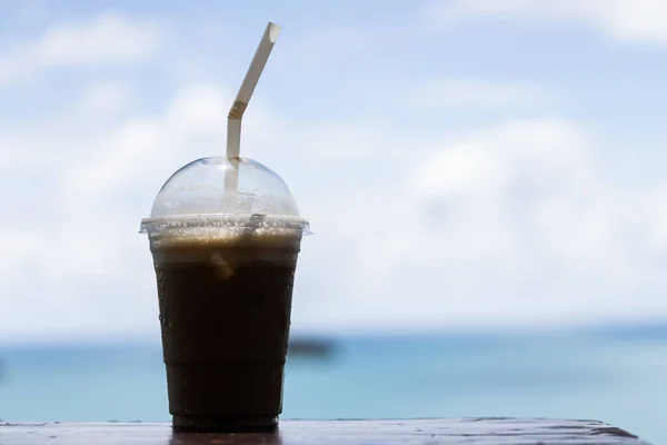 Iced coffee in a plastic cup, perched on a table, with sky and sea backdrop, ideal for background.