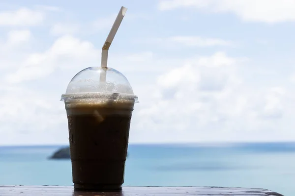 Iced coffee in a plastic cup, perched on a table, with sky and sea backdrop, ideal for background.