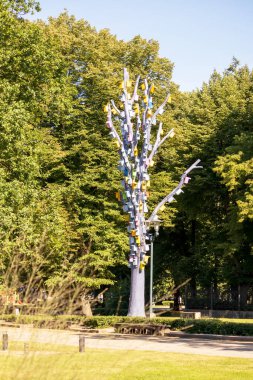 Birbirinin üstüne yığılmış yeşil ağaç zeminli, renkli kuş kafesleriyle dolu gri bir ağaç.