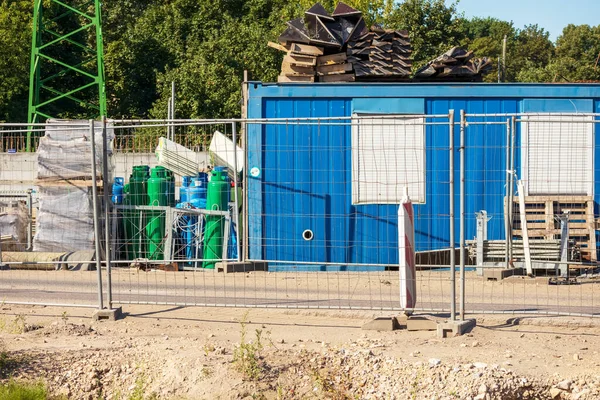 Construction site with a blue metal house and green trees and a metal fence in front of it