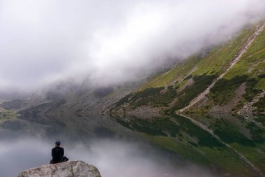 Polonya Tatra Dağları 'nda, gölü ve büyük gri bir kayanın üzerinde oturan bir adam ile huzurlu bir manzara.