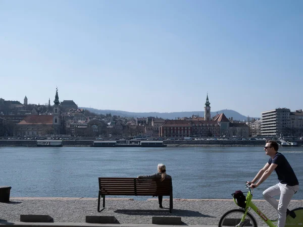 Budapeşte, Macaristan - Mart 2022: Bankta oturan yalnız kadın Tuna Nehri 'ni seyrediyor ve Budapeşte' de bisiklete binen adam, seçici odaklı