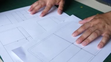 Adam yeşil bir masaya beyaz çarşaf ve çizimler koyuyor. Cüzdan dikmek için desenli çizimler..