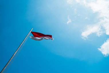 Red and white flag, Indonesia's national flag flying above the pole for independence day in a bright blue sky with white clouds around it