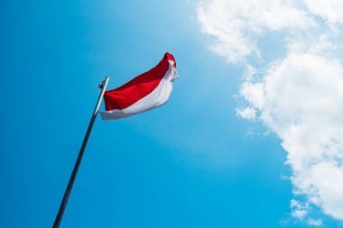 Red and white flag, Indonesia's national flag flying above the pole for independence day in a bright blue sky with white clouds around it