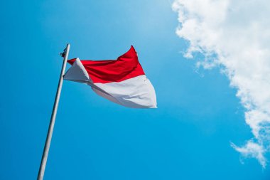 Red and white flag, Indonesia's national flag flying above the pole for independence day in a bright blue sky with white clouds around it