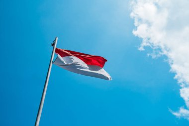 Red and white flag, Indonesia's national flag flying above the pole for independence day in a bright blue sky with white clouds around it