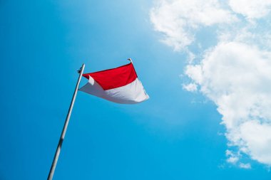 Red and white flag, Indonesia's national flag flying above the pole for independence day in a bright blue sky with white clouds around it