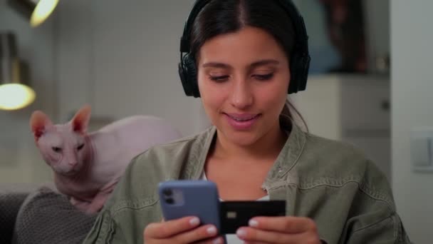 Ein junges Mädchen bezahlt im Internet per Handy. Mädchen sitzt zu Hause auf der Couch mit einer Katze und hört Musik über Kopfhörer — Stockvideo