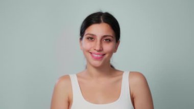 Genç bir esmer kız beyaz bir arka planda yüzünü pamukla ovuyor. Kız yeni bir yüz ifadesi deniyor ve gülümsüyor. Yakın plan.