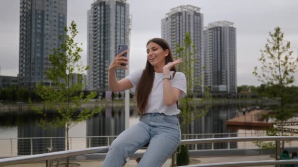 En ung brunett flicka sitter i en park på ett räcke och kommunicerar via videosamtal på telefonen. Flickan har vit t-shirt och jeans. Kommunikation via telefon, videokommunikation, modern värld — Stockvideo