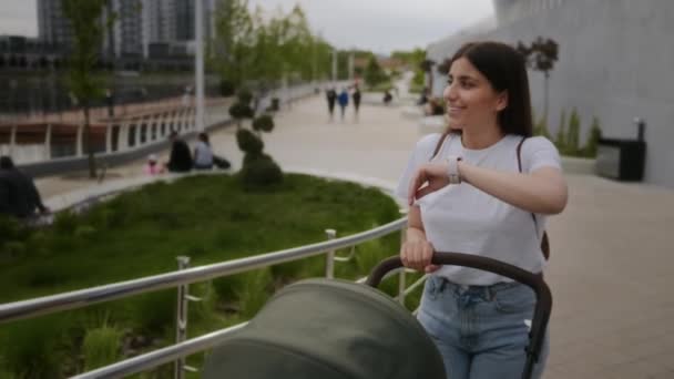 La chica camina con un cochecito en el parque y habla en un reloj inteligente. Mamá sonriente camina con un bebé en un cochecito en la calle y habla a través del reloj. Llamada telefónica, reloj inteligente, comunicación — Vídeo de stock