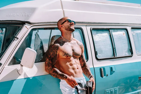 man with tattoo and tattoos leaning against a blue caravan on the beach