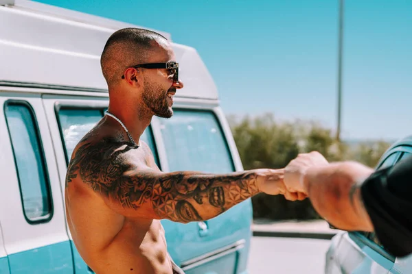 man with tattoos leaning against a blue caravan on the beach