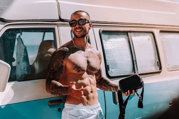 man with tattoo and tattoos leaning against a blue caravan on the beach