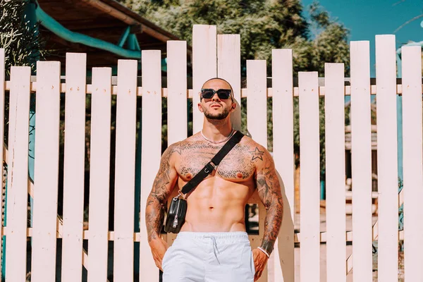 young muscular man with tattoos leaning on a white picket fence on the beach