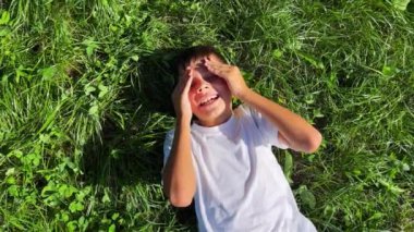 A young boy lies in the grass, smiling, hiding his face.