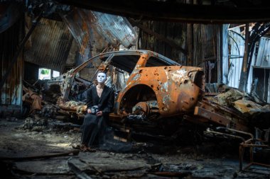 A girl in a black suit and a white mask sits near a burnt-out car from a Russian army shell hitting the house.