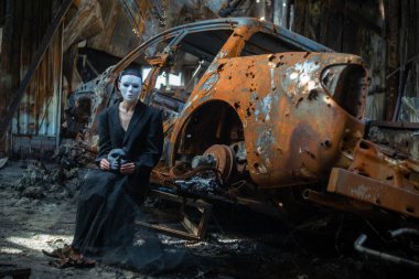 A girl in a black suit and a white mask sits near a burnt-out car from a Russian army shell hitting the house.