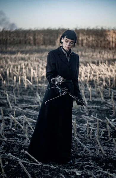 一个穿着时髦黑色夹克和长裙的女孩站在田野中央 手里拿着一朵枯萎的向日葵 — 图库照片