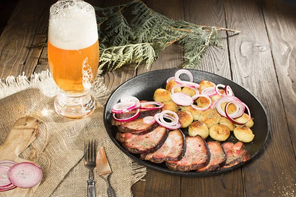 lunch of fried ham with potatoes and onions in a frying pan next to a glass of beer on a rustic wooden table
