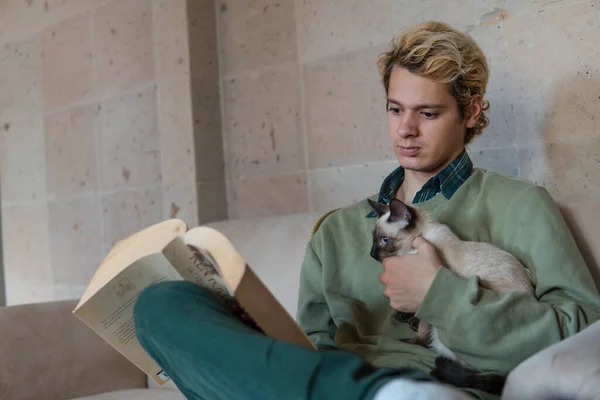 Blond man is reading a book on the sofa in the living room of his house while carrying his cat which reads the book at the same time