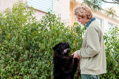 Sarışın bir çocuk ve büyük siyah bir köpek sahibinin yardımıyla dışarıda oynuyorlar.
