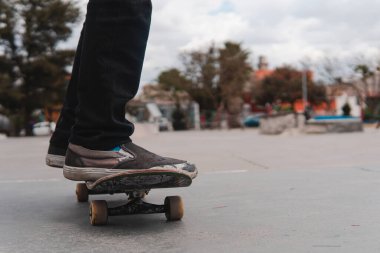 The feet of a boy with jeans and sneakers on a skateboard with the park in the background