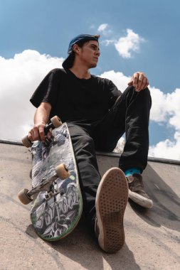 Ön planda kaykayı ve tenis ayakkabılarıyla siyah giyinmiş bir çocuk. Arka planda gökyüzü var. Parktaki bir rampada oturuyor.