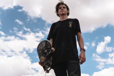 Genç bir patenci sörf tahtasını tutarken arka planda gökyüzü ile poz veriyor. Çocuk kotla siyah giyiyor..